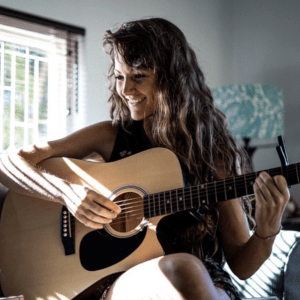 Interview Music Producer Kirsten Hunneyball, student at Abbey Road Institute Johannesburg sitting on bed playing guitar, smiling with long curly hair